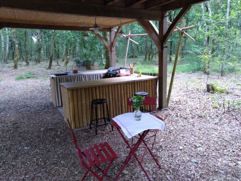 Cabane en bois avec bar extérieur et table rouge, entourée de verdure.