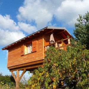 Cabane en bois perchée, entourée de verdure sous un ciel bleu parsemé de nuages.