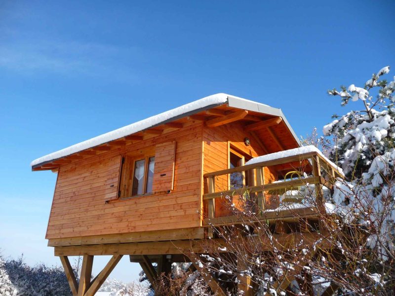 Cabane en bois sur pilotis, entourée de neige, sous un ciel bleu.