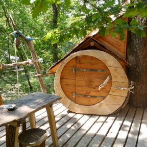 Cabane perchée en bois avec un accès original à une porte ronde, entourée darbres.