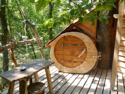 Cabane perchée en bois avec un accès original à une porte ronde, entourée darbres.