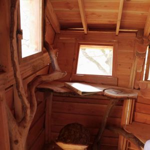 Cabane en bois dans les arbres, avec une table en bois brut et de petites fenêtres.