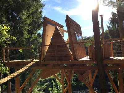 Cabane perchée en bois avec toit en ardoise, entourée darbres verdoyants.