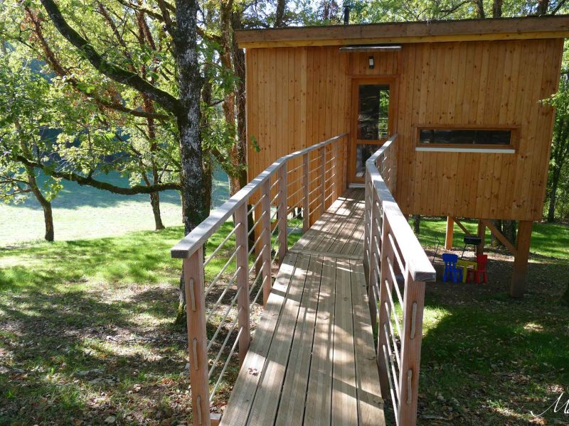 Cabane en bois sur pilotis, accessible par une passerelle en bois, entourée darbres.