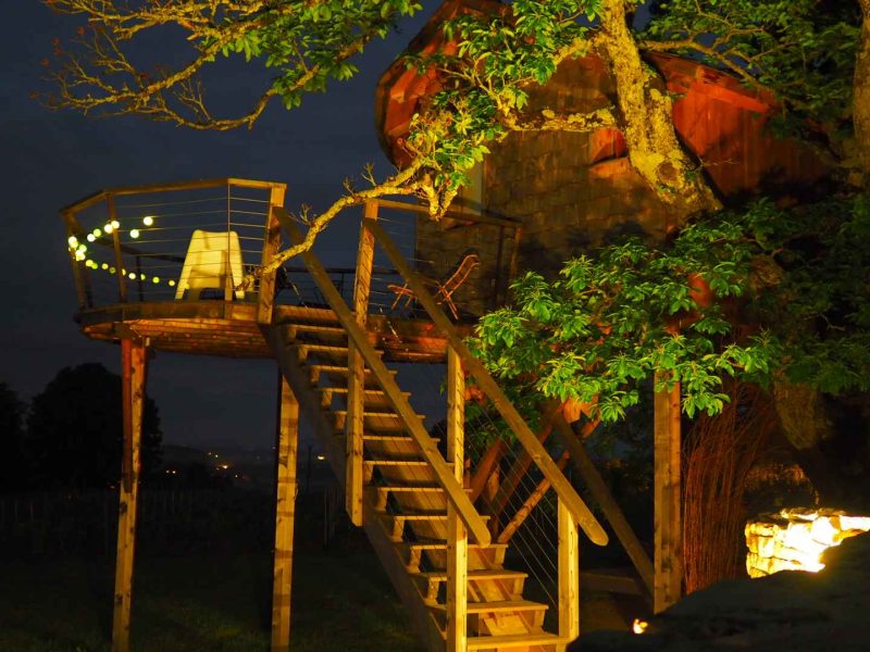 Cabane dans les arbres illuminée, avec une terrasse et des escaliers en bois.