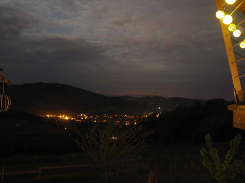 Cabane perchée avec guirlande lumineuse, vue nocturne sur un village illuminé.
