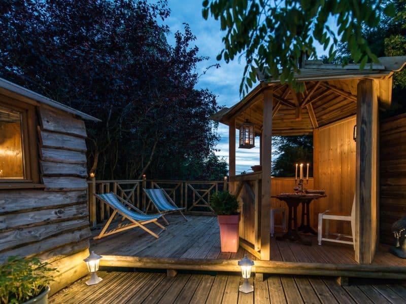 Cabane en bois avec terrasse éclairée, chaises longues et ambiance chaleureuse.
