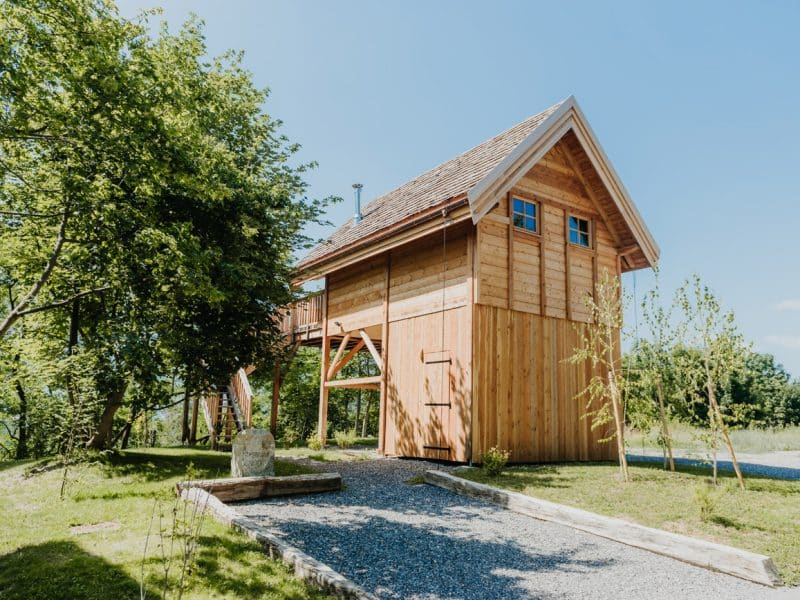Cabane en bois perchée, entourée darbres et dun chemin en gravier.