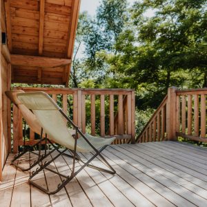 Cabane en bois avec terrasse ensoleillée et fauteuil relaxant, entourée de verdure.