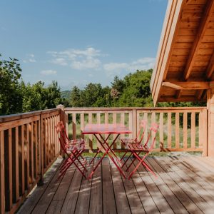 Cabane en bois avec terrasse, table et chaises rouges, vue sur la nature.