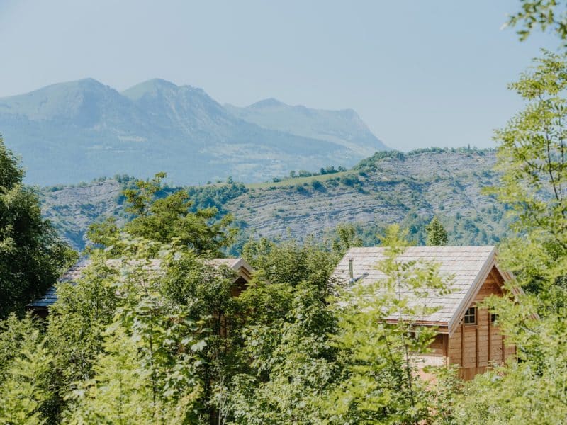 Chalets en bois entourés de verdure, avec montagnes en arrière-plan.
