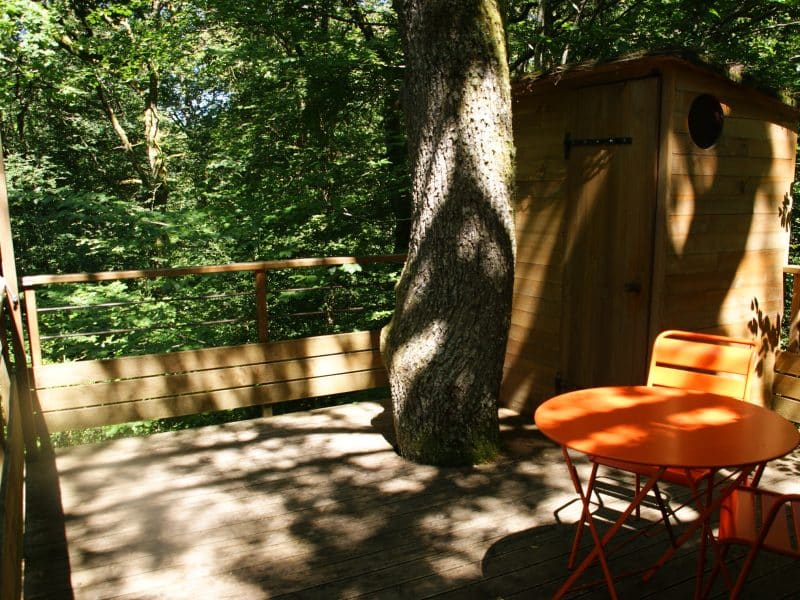 Cabane dans les arbres avec terrasse en bois et mobilier orange, entourée de verdure.