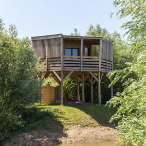 Cabane sur pilotis en bois, entourée de verdure, avec chaises longues au bord de leau.