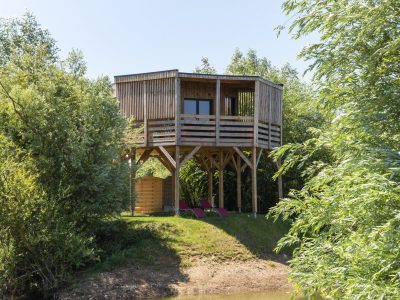 Cabane sur pilotis en bois, entourée de verdure, avec chaises longues au bord de leau.