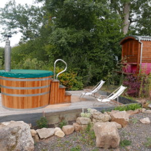 Hébergement insolite en roulotte avec jacuzzi en bois et transats dans un cadre verdoyant.