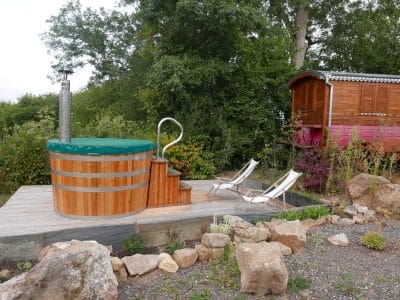 Hébergement insolite en roulotte avec jacuzzi en bois et transats dans un cadre verdoyant.