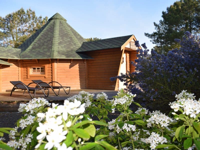 Chalet en bois avec toit en pointe, entouré de fleurs blanches et violettes.
