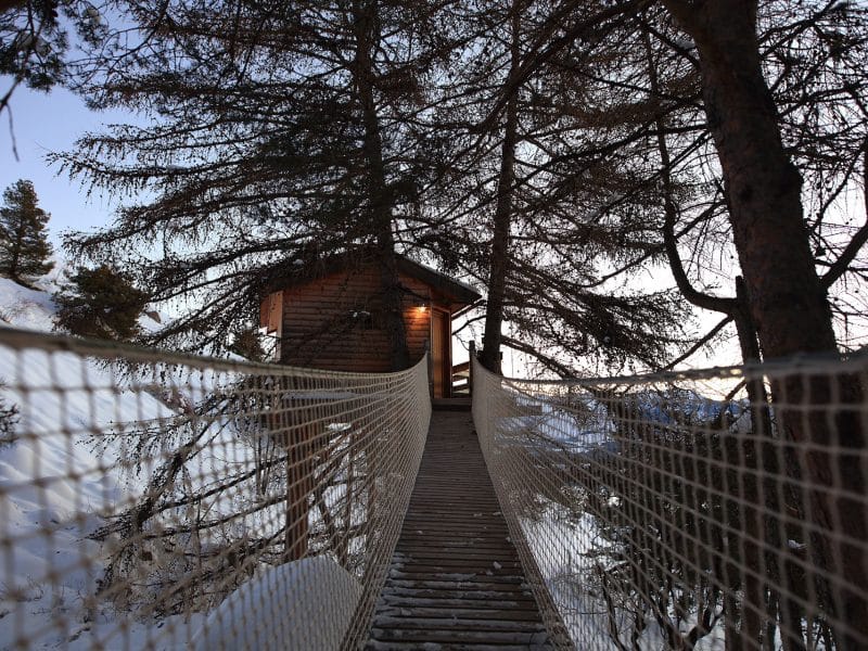 Cabane perchée dans les arbres, accessible par un pont suspendu, au cœur des Alpes.