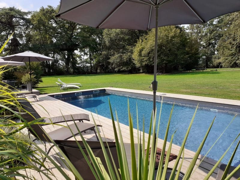 Villa avec piscine, chaises longues et parasols entourés dun jardin verdoyant.