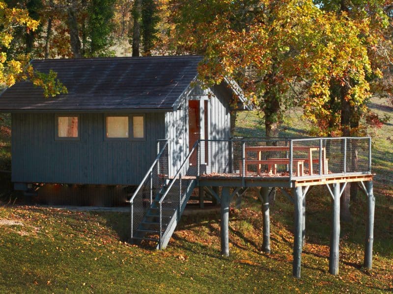 Cabane sur pilotis entourée darbres, avec une terrasse en bois accueillante.