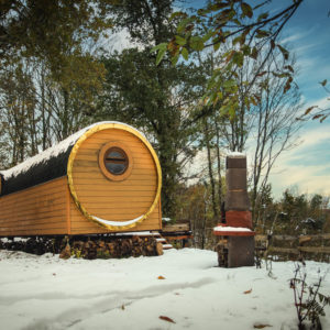 Hébergement insolite en forme de tonneau, entouré de neige et de nature verdoyante.