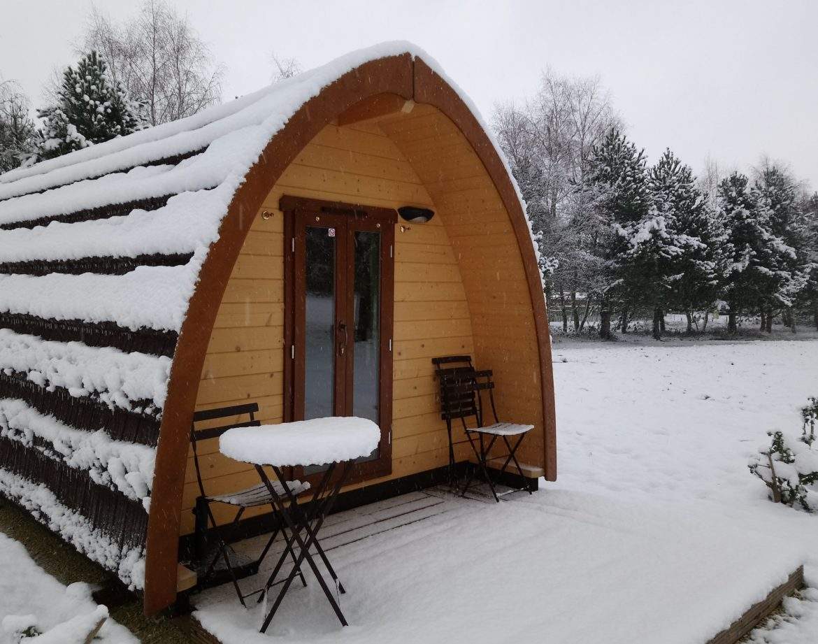 Cabane en bois cosy sous la neige, avec terrasse et chaises en extérieur.