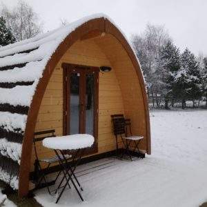 Cabane en bois cosy sous la neige, avec terrasse et chaises en extérieur.