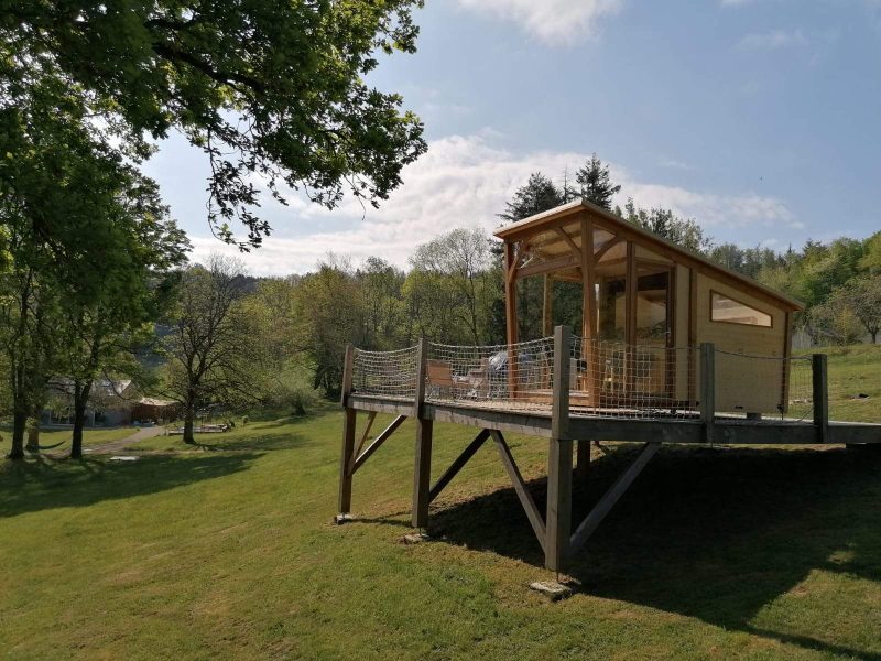 Cabane sur pilotis en bois, entourée de verdure et baignée de lumière naturelle.