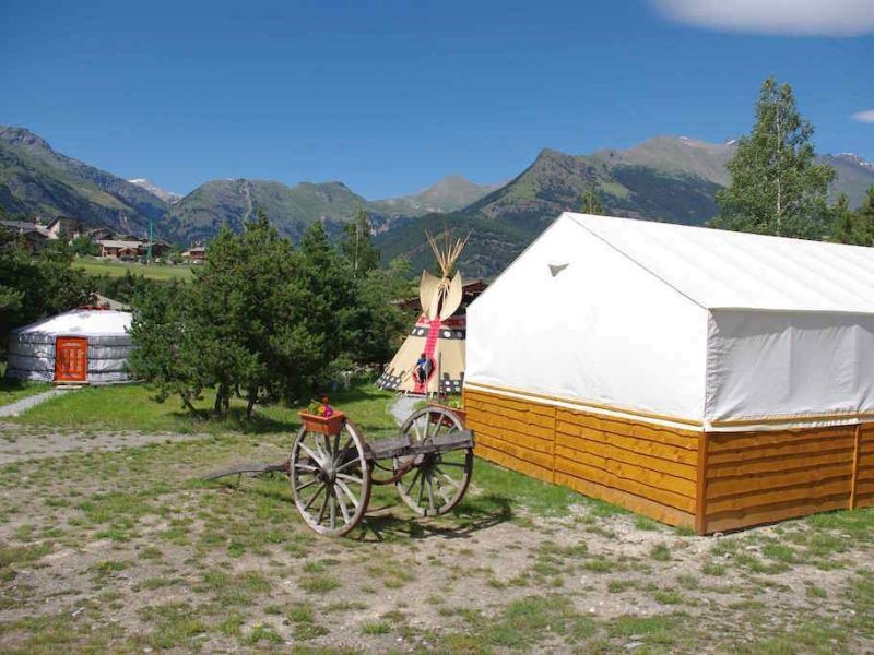 Tente de glamping avec vue sur les montagnes, entourée de verdure et dune charrette en bois.