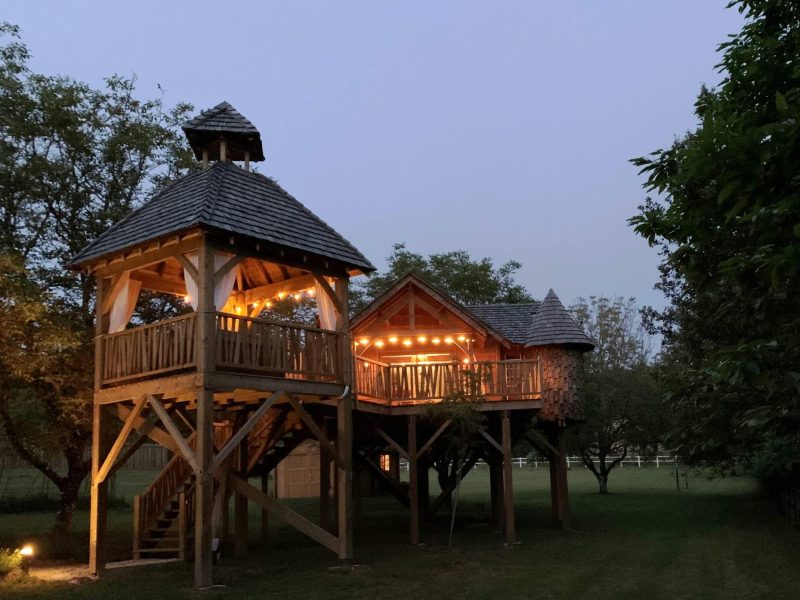 Cabane perchée en bois, illuminée, entourée darbres verdoyants au crépuscule.