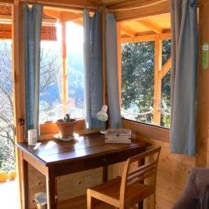 Cabane en bois lumineuse avec vue sur la nature, bureau en bois et rideaux bleus.