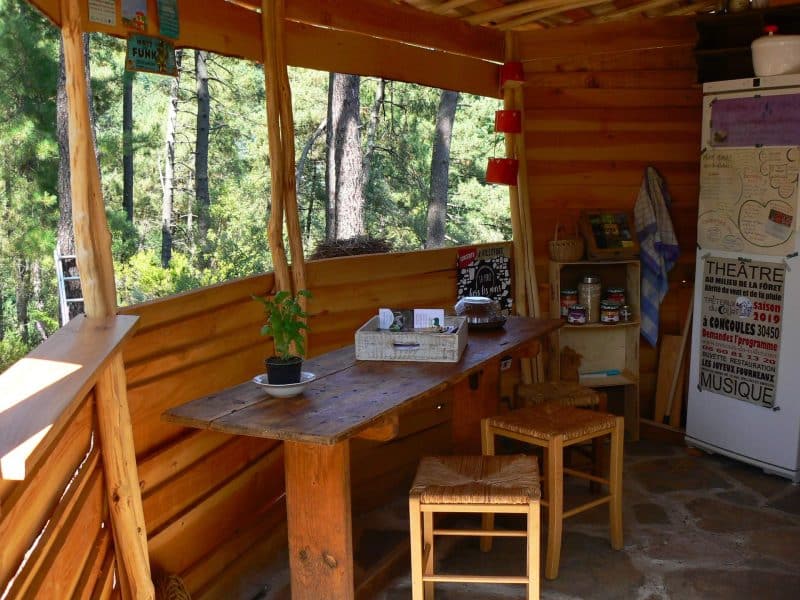 Cabane en bois avec une table en bois et des chaises, entourée de verdure.