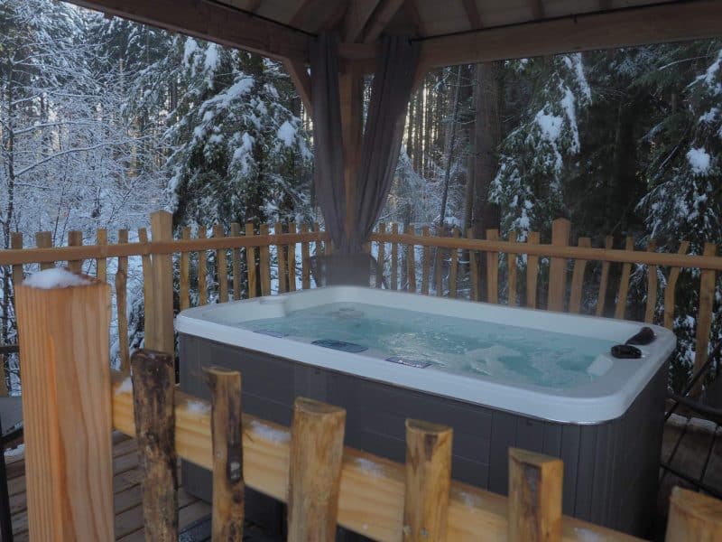Cabane en bois avec jacuzzi, entourée de neige et de forêt verdoyante.
