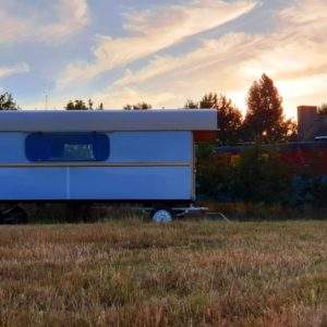 Caravane insolite au coucher de soleil, entourée de verdure et de champs dorés.