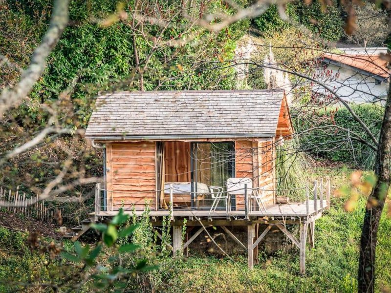 Cabane en bois sur pilotis, entourée de verdure, avec terrasse et chaises.