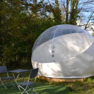Hébergement insolite en bulle transparente, entouré de verdure, avec table et chaises.