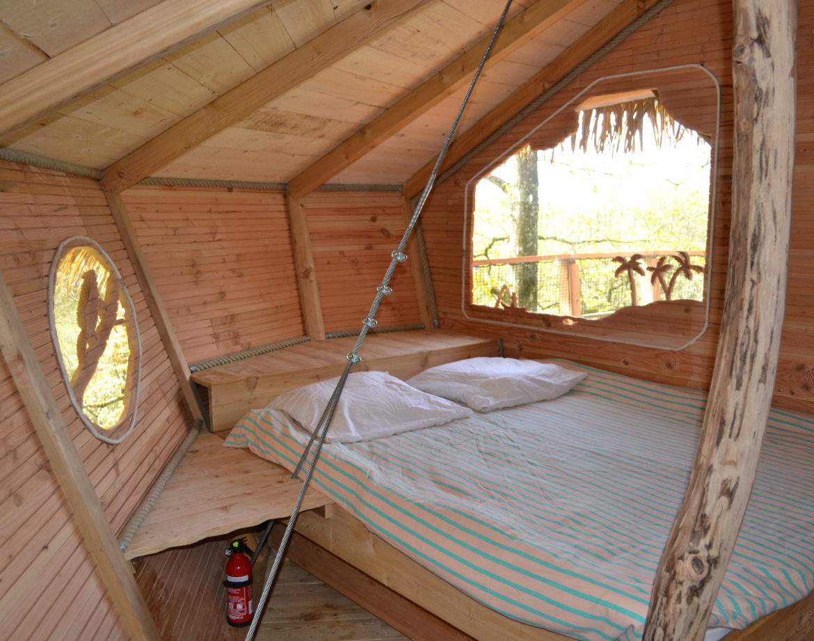 Cabane en bois avec lit douillet et fenêtre ronde, située dans la nature.