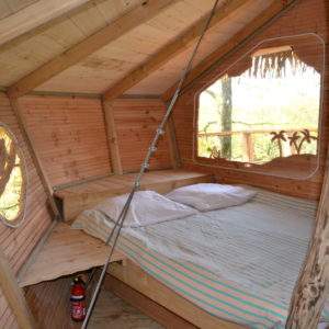 Cabane en bois avec lit douillet et fenêtre ronde, située dans la nature.
