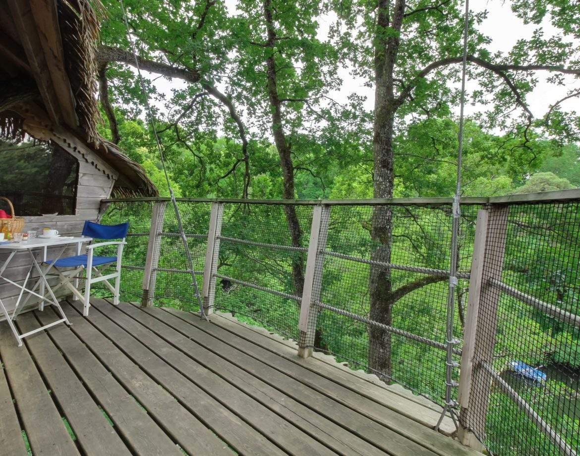 Cabane perchée dans les arbres avec terrasse en bois et vue sur la nature verdoyante.