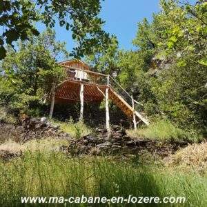 Cabane perchée en bois dans la nature, entourée de verdure et de collines.