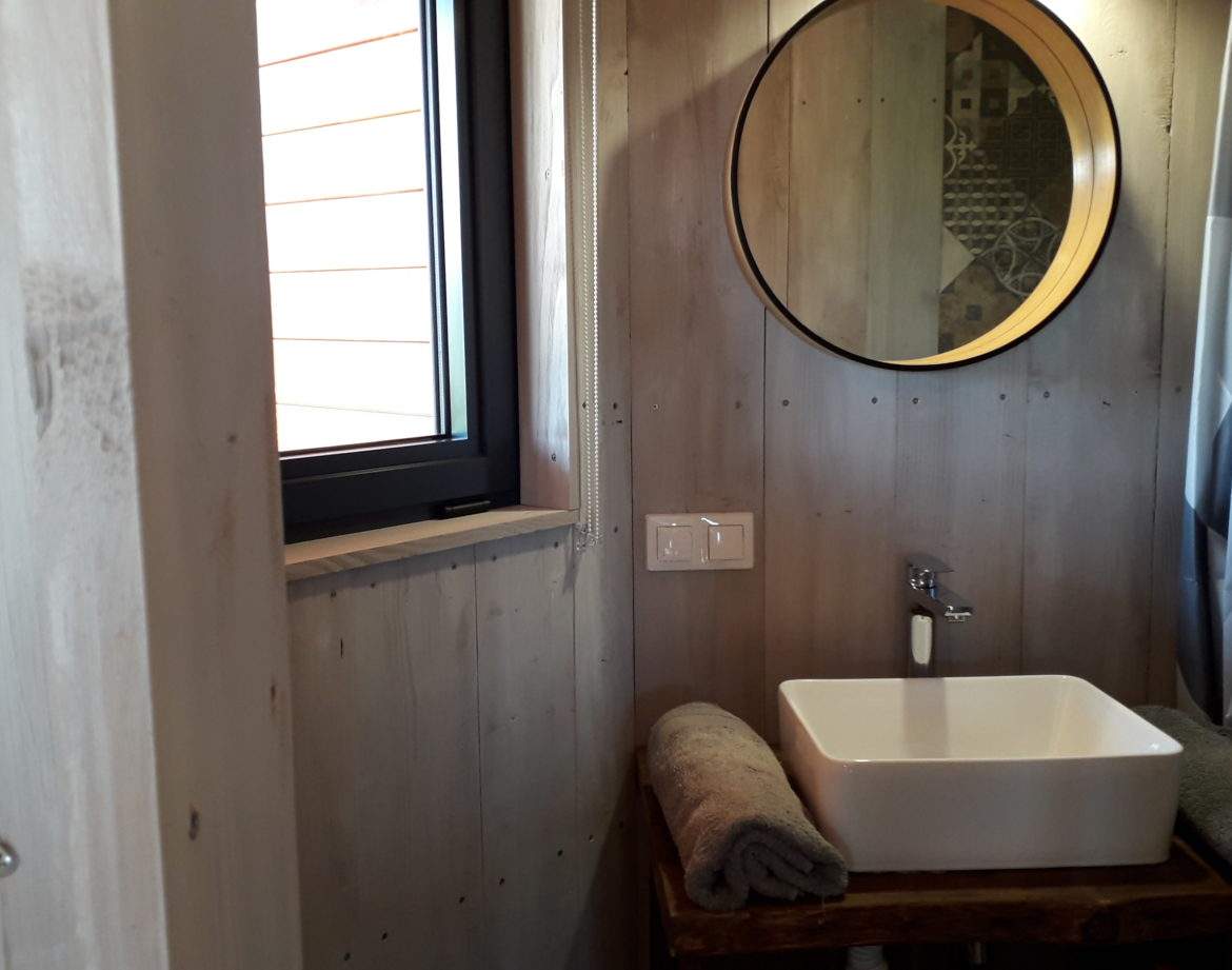 Salle de bain moderne dans un hébergement insolite en bois, avec miroir rond et lumière naturelle.