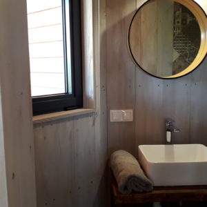 Salle de bain moderne dans un hébergement insolite en bois, avec miroir rond et lumière naturelle.