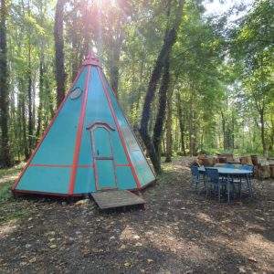 Tipi coloré en pleine forêt, entouré de verdure et dune table en bois.