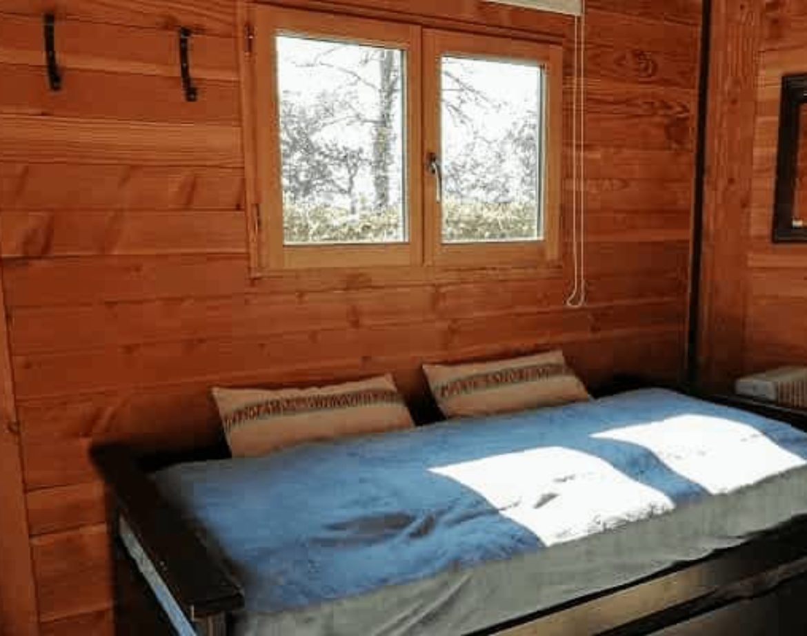 Cabane en bois avec lit confortable et grandes fenêtres lumineuses.