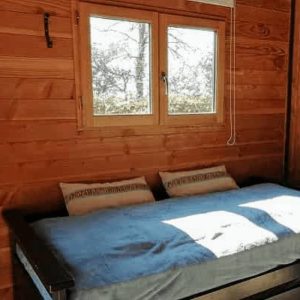 Cabane en bois avec lit confortable et grandes fenêtres lumineuses.