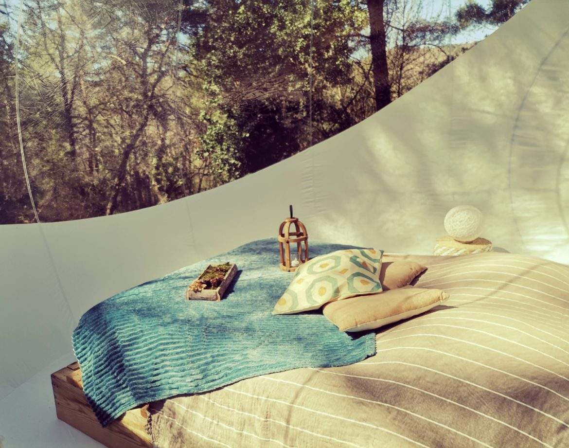 Hébergement insolite en bulle, lit douillet avec coussins colorés et vue sur la nature.
