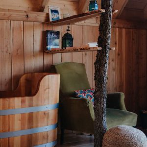 Cabane en bois avec baignoire en bois et fauteuil confortable, ambiance chaleureuse.