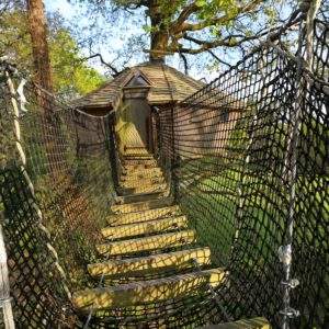 Cabane perchée en Bretagne, accessible par un pont en filet, entourée de verdure.
