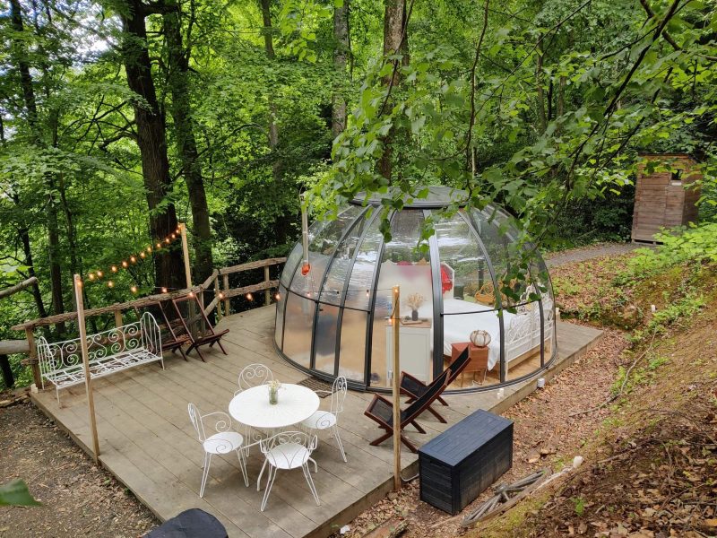 Dôme transparent en pleine forêt, avec terrasse et mobilier en bois. Idéal pour se ressourcer.
