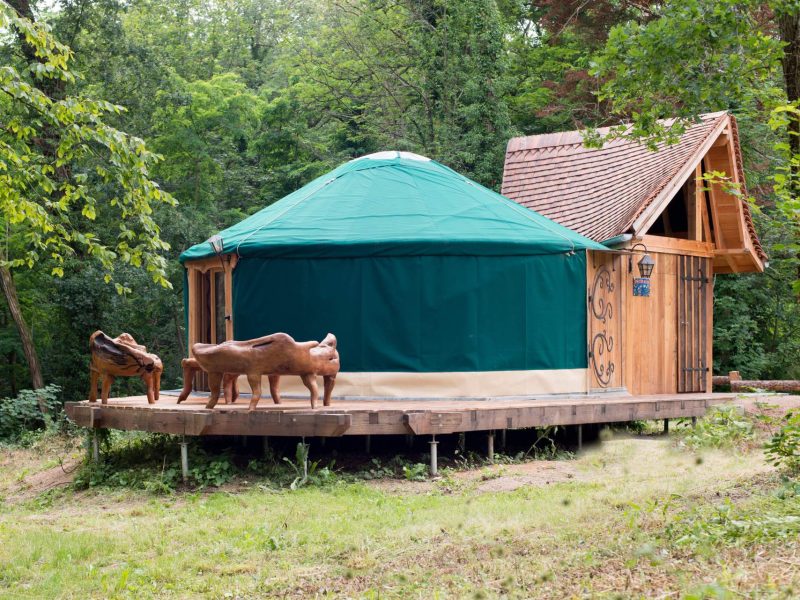 Yourte en bois entourée darbres, avec sculptures en bois sur la terrasse.